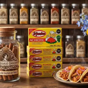 Cajas de achiote de 100g marca el yucateco, sobre mesa de madera, acompañadas de un frasco de canela en vara y tacos de cochinita pibil, al fondo un estante de madera con frascos de especias de Lady Regaliz Grocery.